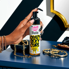 Hand holding a bottle of 'Crazy Sexy Curl' foam with jewelry on a reflective surface